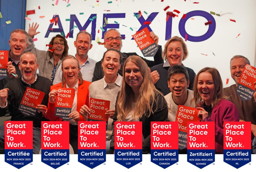 Un groupe de personnes célébrant avec de grands sourires et tenant des plaques de certification « Great Place to Work » pour divers pays, notamment la France, la Belgique, le Portugal, le Luxembourg, le Canada, la Suisse et les Pays-Bas. Des confettis tombent en arrière-plan, et le mot « AMEXIO » est partiellement visible derrière eux.